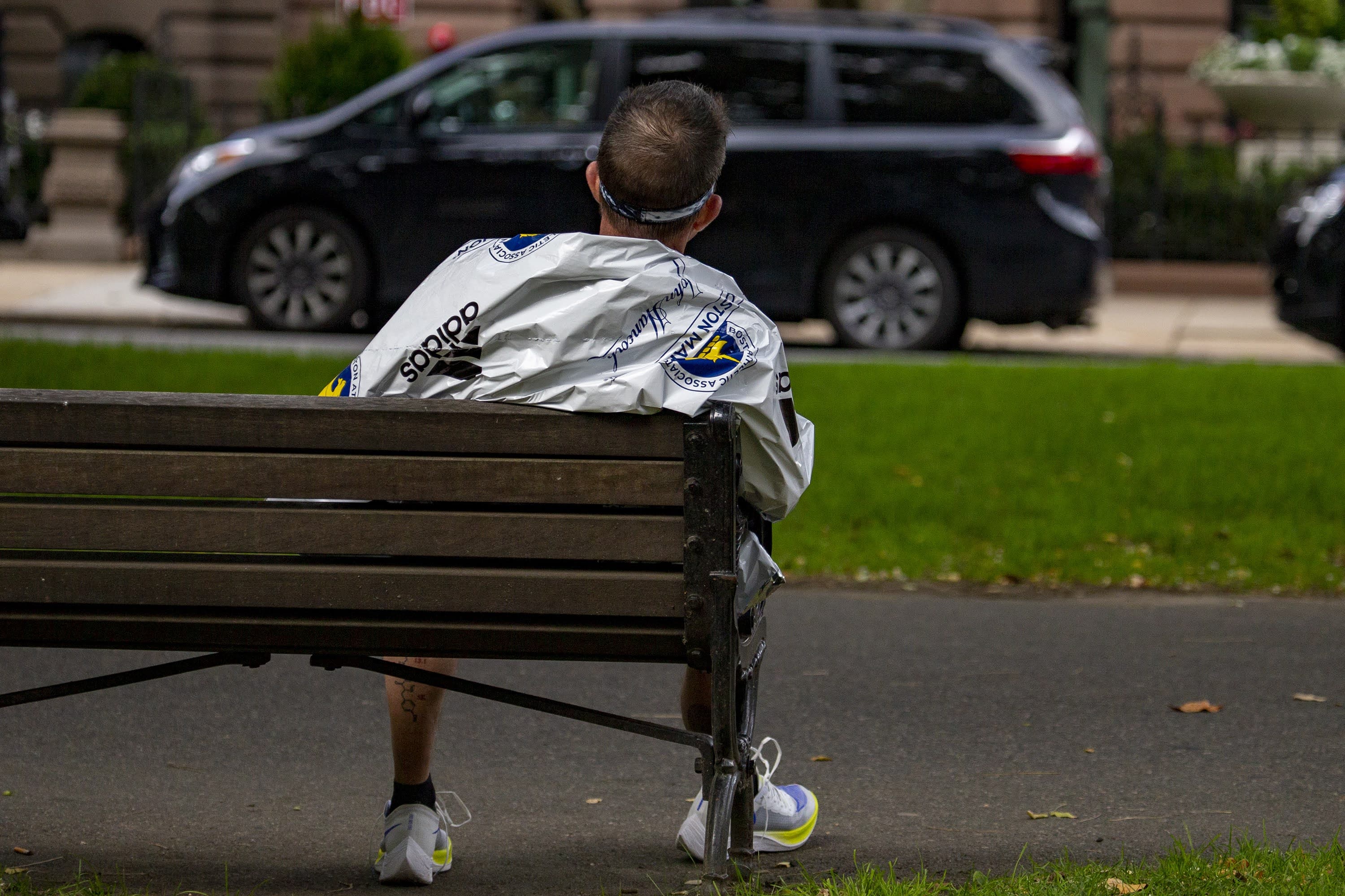 A marathon participant wrapped in a heatsheet blanket stits exhausted on a bench on the Commonwealth Mall after finishing the Boston Marathon. (Jesse Costa/WBUR)
