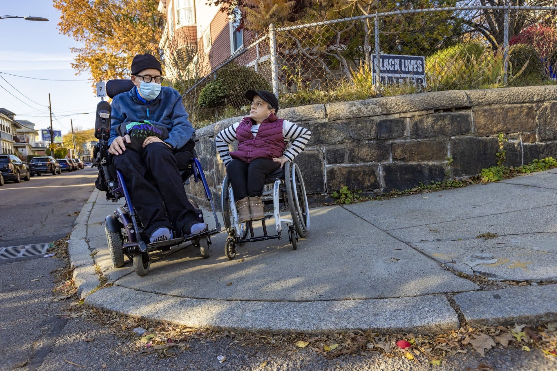Michael Muehe and Colleen Flanagan sit at the edge of a sidewalk with no curb ramp on the corner of Boylston Street and Belmore Terrace in Jamaica Plain. (Jesse Costa/WBUR)