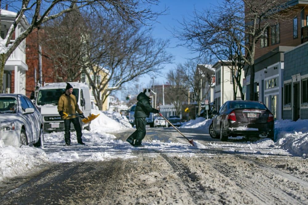 Cameron Minor watches as Rhianna Del Bene pushes a shvel ful of snow across Cross street in Somerville. (Jesse Costa/WBUR)