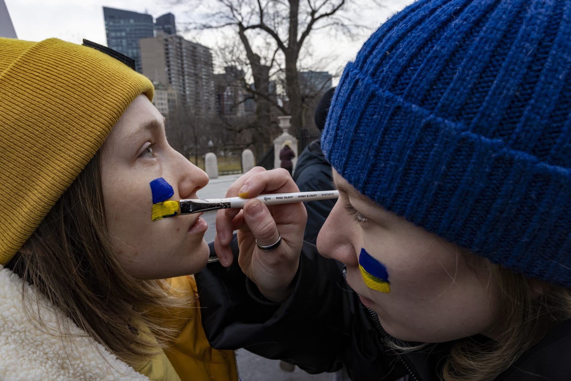 Anna Muranova paints the Ukrainian flag on the face of Tetiana Litus during the State House rally. (WBUR)