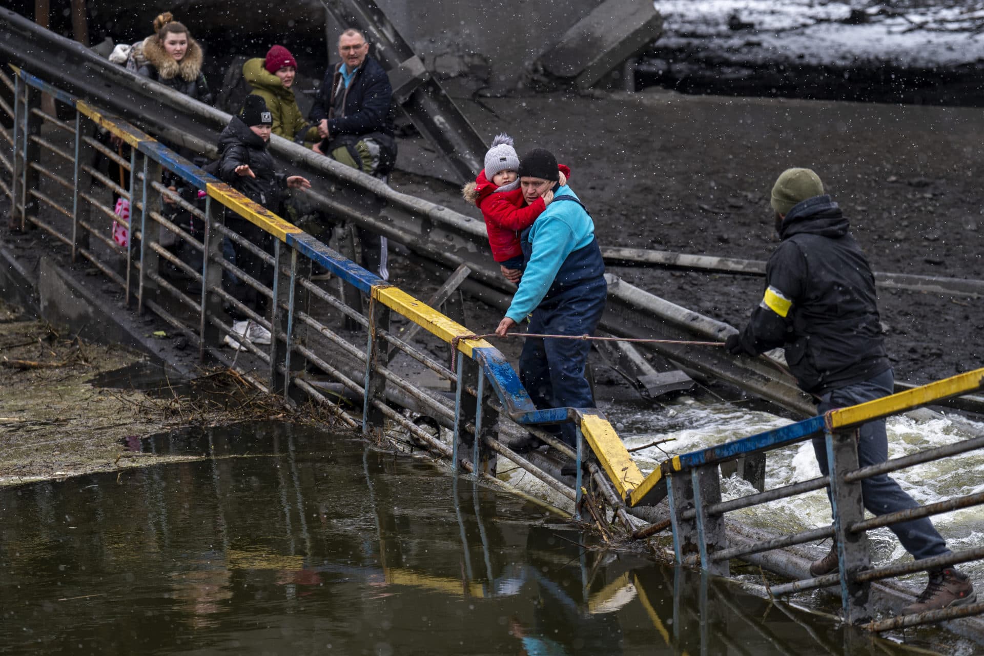 A neighbor carries a child as he helps a fleeing family across a destroyed bridge on the outskirts of Kyiv on Wednesday, March 2. Russian forces have escalated their attacks on crowded cities in what Ukraine's leader called a blatant campaign of terror. (Emilio Morenatti/AP)