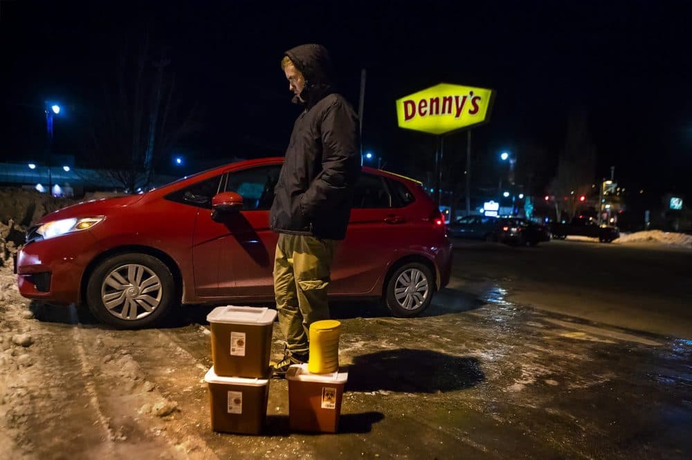 In this 2019 file photo, Jesse Harvey stands in the parking lot of a Denny's in Auburn, Maine, behind four containers filled with used needles collected from area drug users. (Jesse Costa/WBUR)