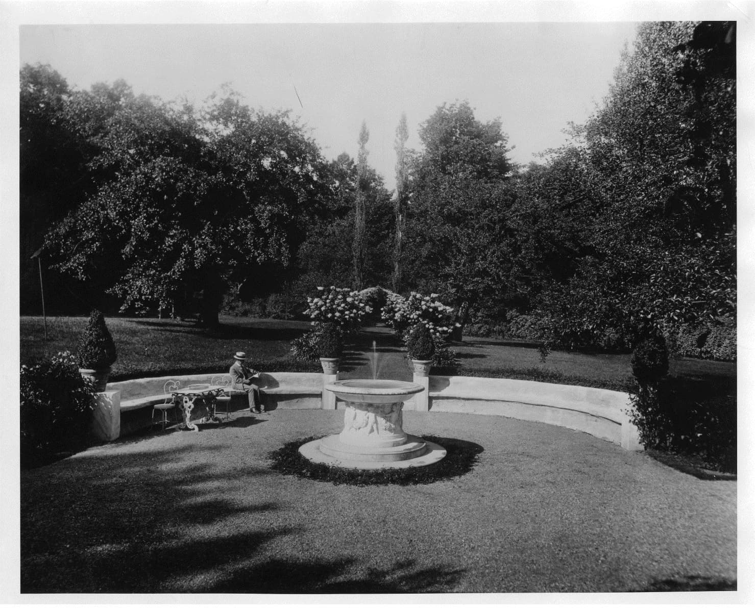 A black and white photo of a rounded garden with American sculptor Daniel Chester French seated on the left side. A fountain at the center. Lots of greenery in the background. The photo is courtesy of the Chesterwood Archives.