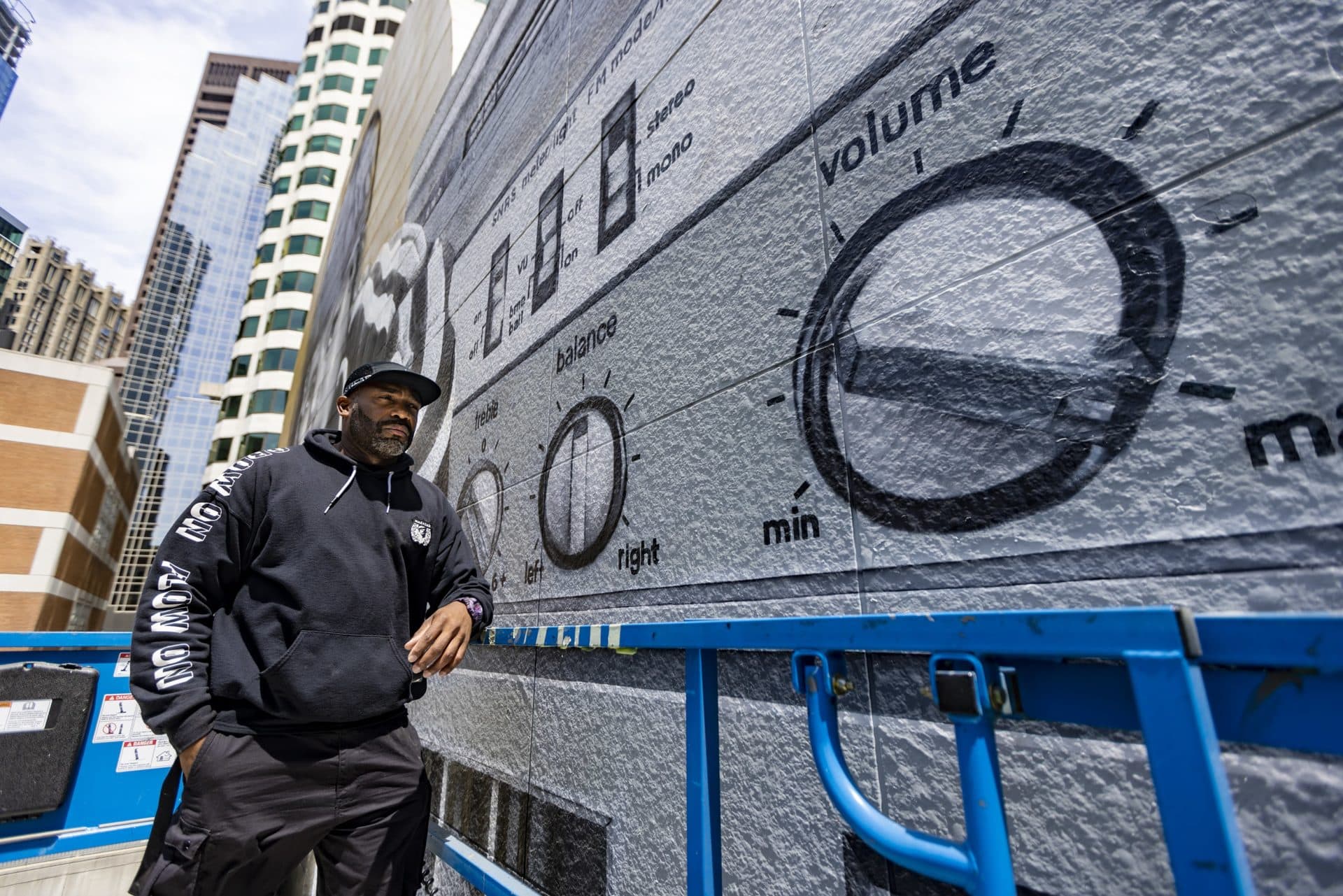 Muralist Rob “ProblaK” Gibbs standing on a scissor lift near the painted control knobs of a boombox in his work “Breathe Life Together,” the next mural to be featured on the façade of the Dewey Square Tunnel Air Intake Structure on the Rose Kennedy Greenway. (Jesse Costa/WBUR)