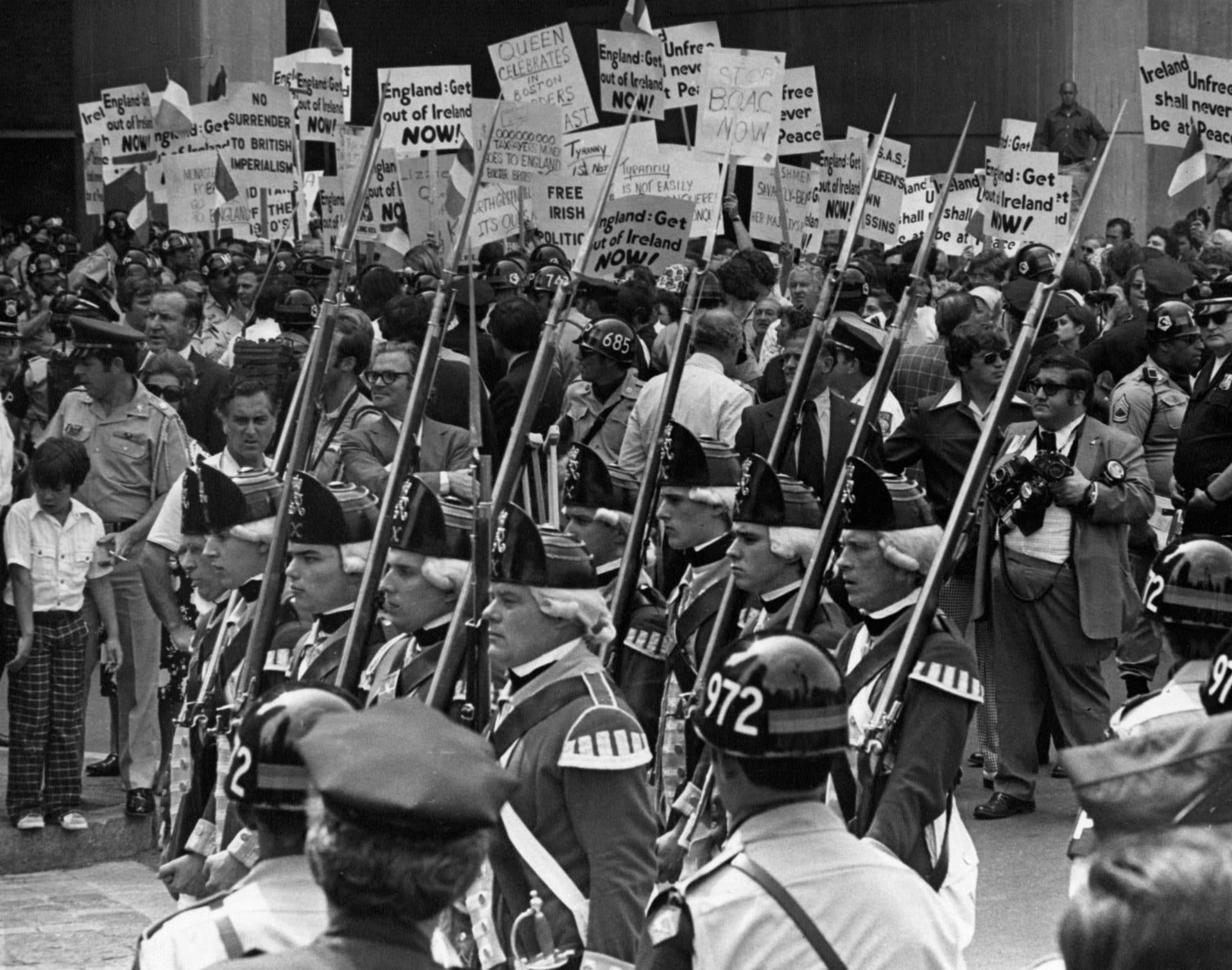 Troops passing in review at the Samuel Adams statue during Queen Elizabeth II's visit. Protesters outside Boston City Hall hold up signs in the background. (Charles Dixon/The Boston Globe via Getty Images)