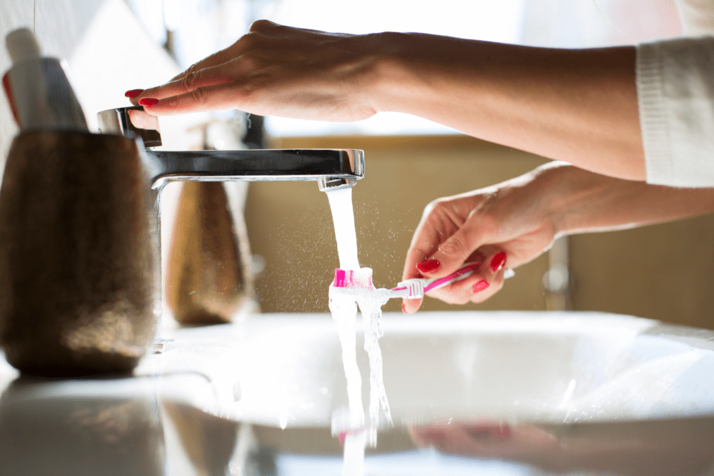 A person holds a toothbrush under running water from the faucet.