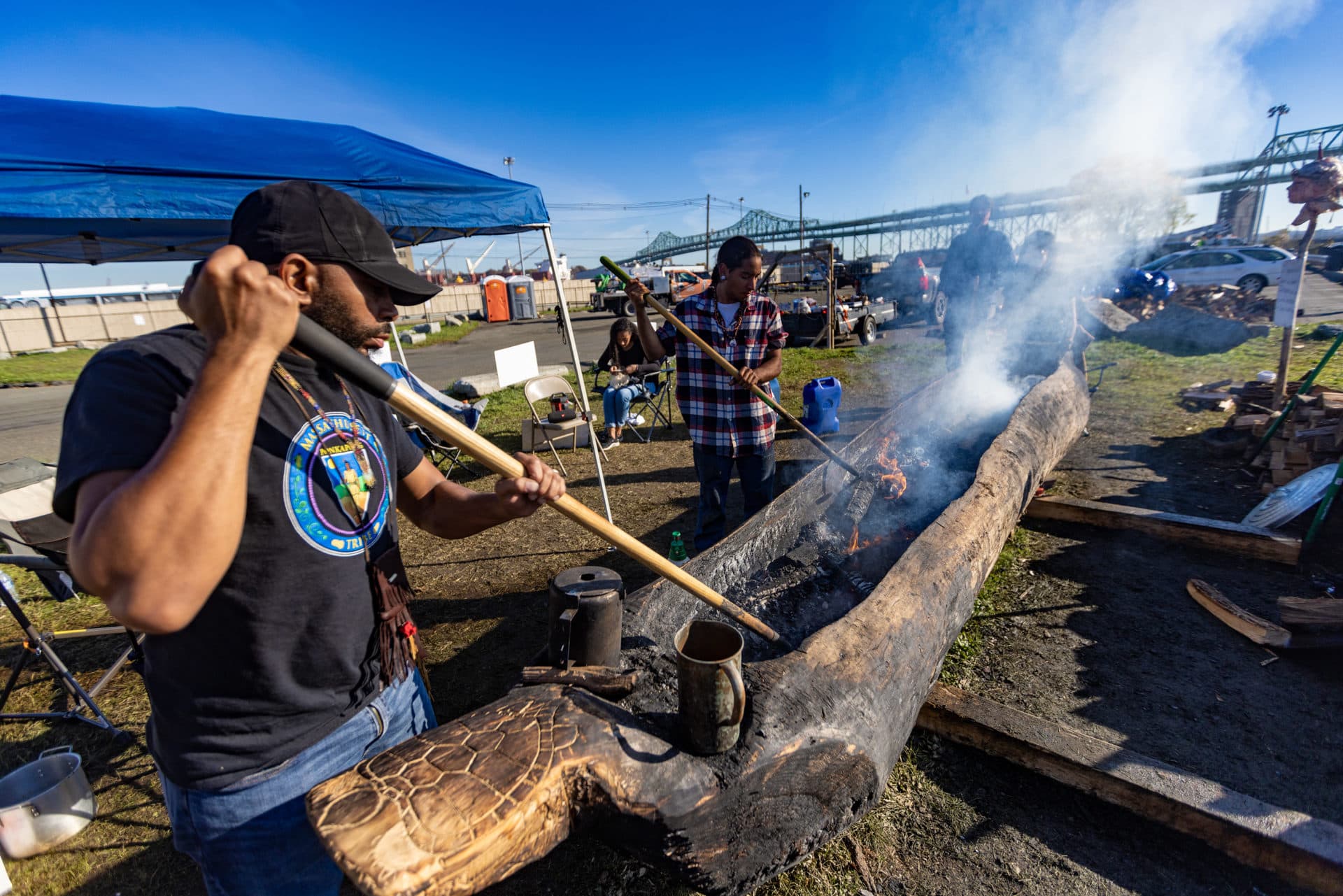 Thomas Green, Indigenous artist and educator of the Massachusett Tribe at Ponkapoag, left, rakes hot coals while burning a traditional mishoon. (Jesse Costa/WBUR)