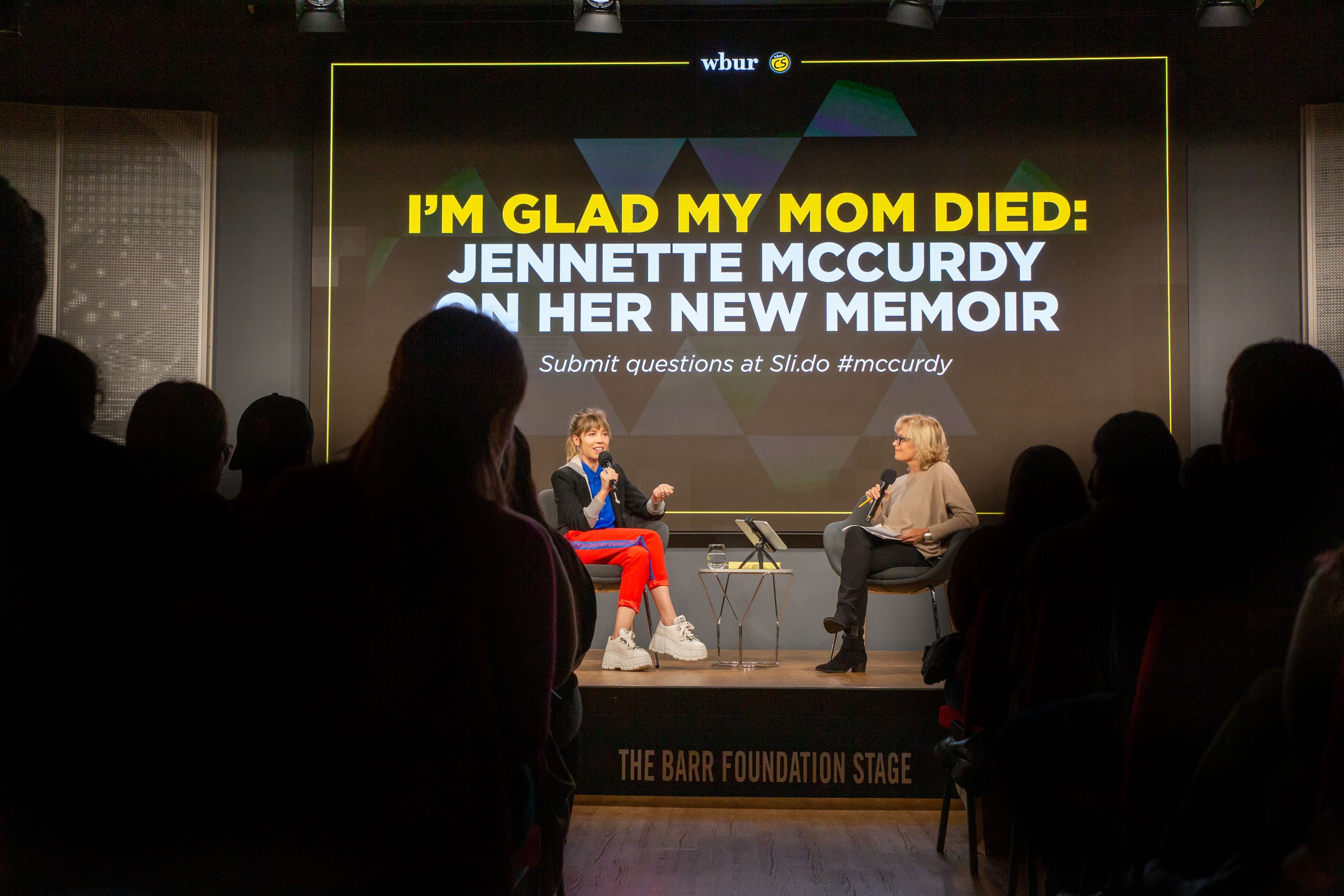 Jennette McCurdy is shown wearing red pants and large white shoes with a black jacket while sitting next to Robin Young who is wearing black pants and a cream-colored sweater.