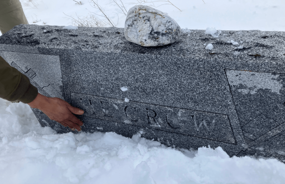 Michael He Crow wipes snow away from the gravestone of his great grandmother and great grandfather, Winona and Jackson He Crow, in Oglala, South Dakota, on Dec. 27, 2022. At age 9, Jackson He Crow not only survived the massacre at Wounded Knee in 1890 by running and hiding, he also rescued his mother who had been shot by the U.S. Cavalry. (Nancy Eve Cohen/NEPM)