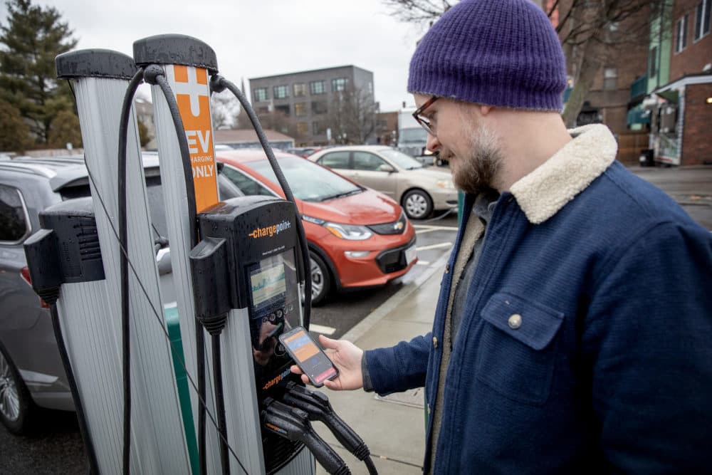 Wesley Sanders uses his phone to connect his car to a charger in Jamaica Plain. He wears a blue coat and a purple wool hat. (Credit: Robin Lubbock/WBUR)