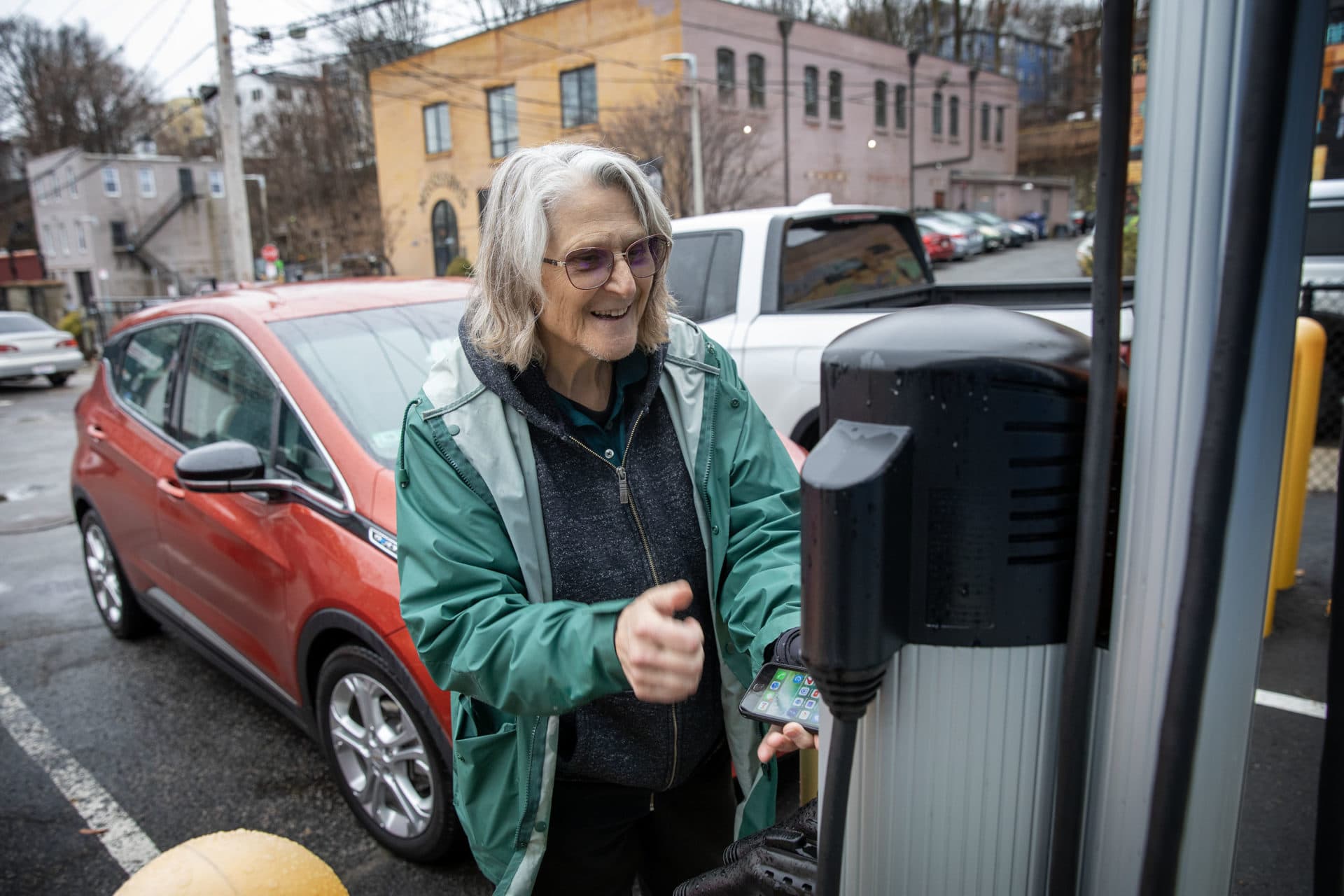Loie Hayes wears a green rain coat and glasses, she's a white woman and has white hair. Hayes uses her phone to connect with a charging station in Boston. She doesn't have a driveway or garage and relies on public chargers in Mission Hill. (Credit: Robin Lubbock/WBUR)