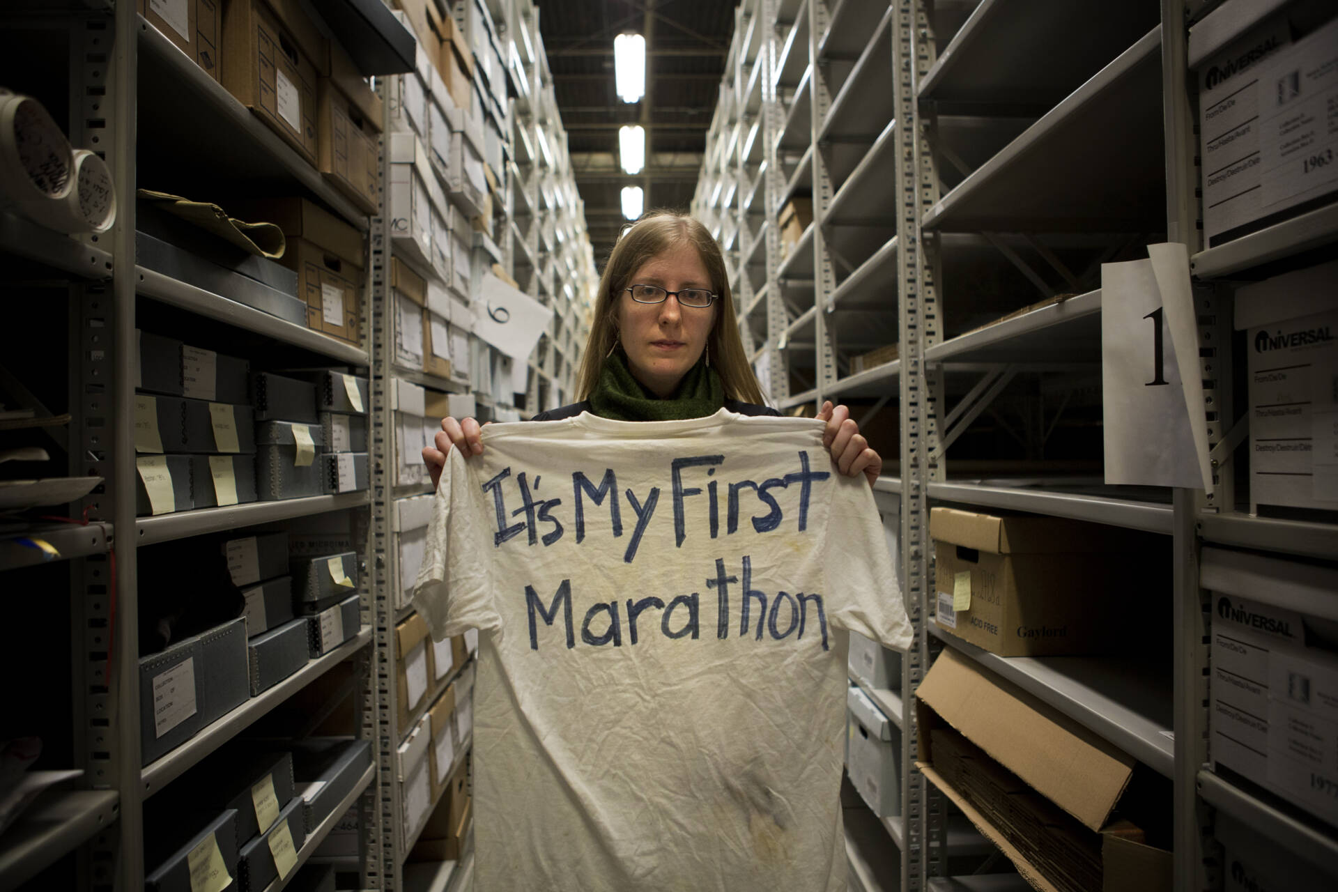 City archivist Marta Crilly holds a &quot;It's My First Marathon&quot; t-shirt inside a cavernous, temperature-controlled warehouse in West Roxbury holding dozens of boxes on its towering shelves that contain items from a makeshift memorial at Copley Square after the bombings. (Jesse Costa/WBUR)