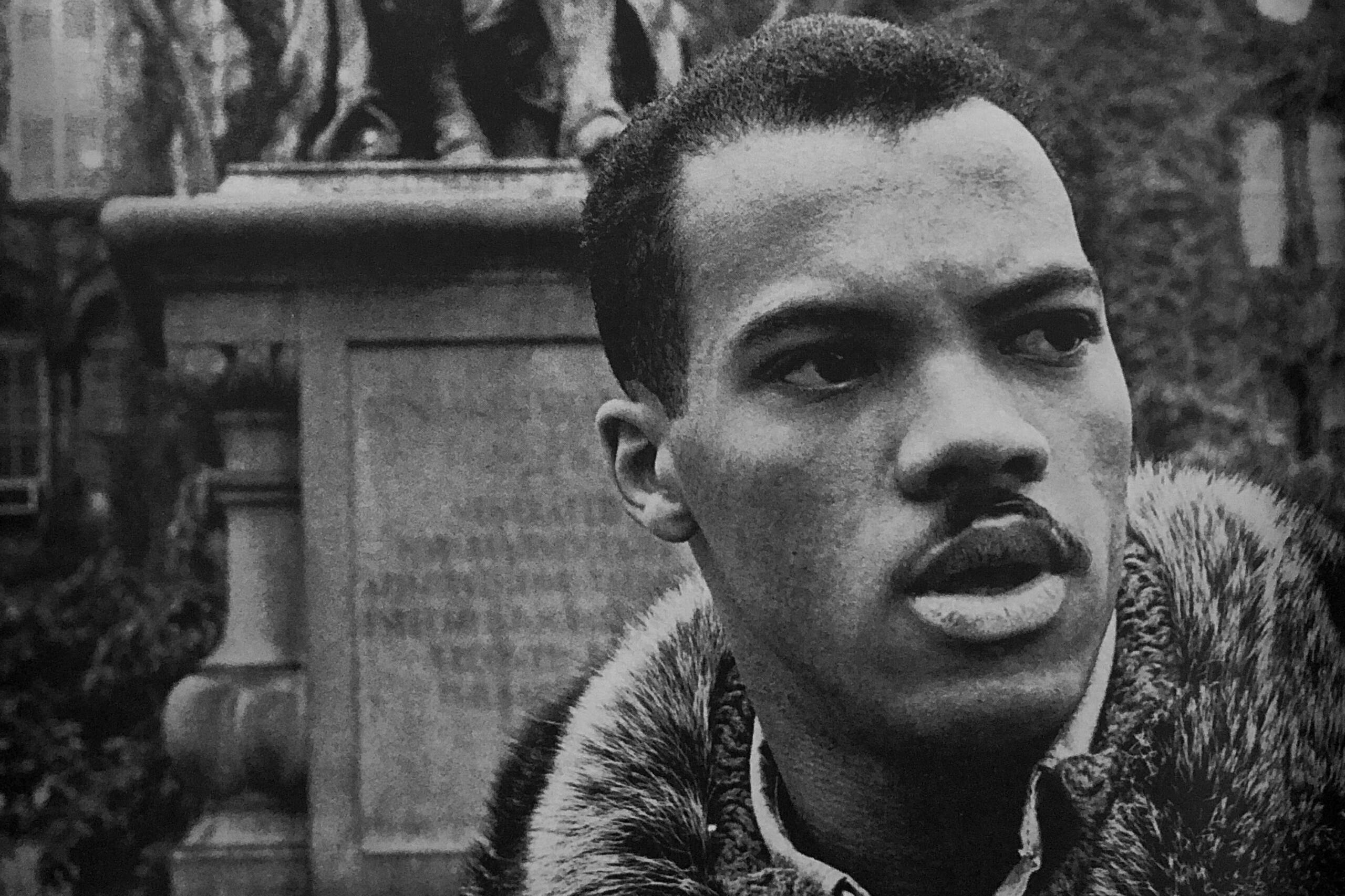 A black and white image of a Black man in a button down shirt and fur-collared jacket, standing in front of a statue. (Jim Hansen/Look Magazine)