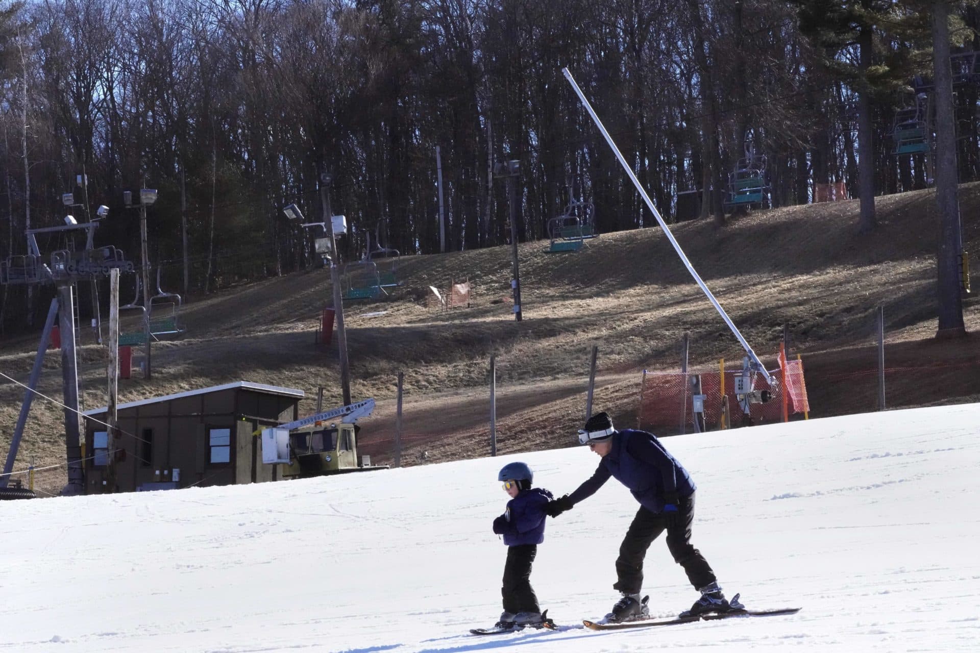 A child learns to ski on the beginners slope, adjacent to snowless snowboarding park, at rear, at the Ski Bradford ski area, Tuesday, Feb. 14, 2023, in in Bradford, Mass. For much of the Eastern United States, the winter of 2023 has been a bust. Snow totals are far below average from Boston to Philadelphia in 2023 and warmer temperatures have often resulted in more spring-like days than blizzard-like conditions. (AP Photo/Charles Krupa)