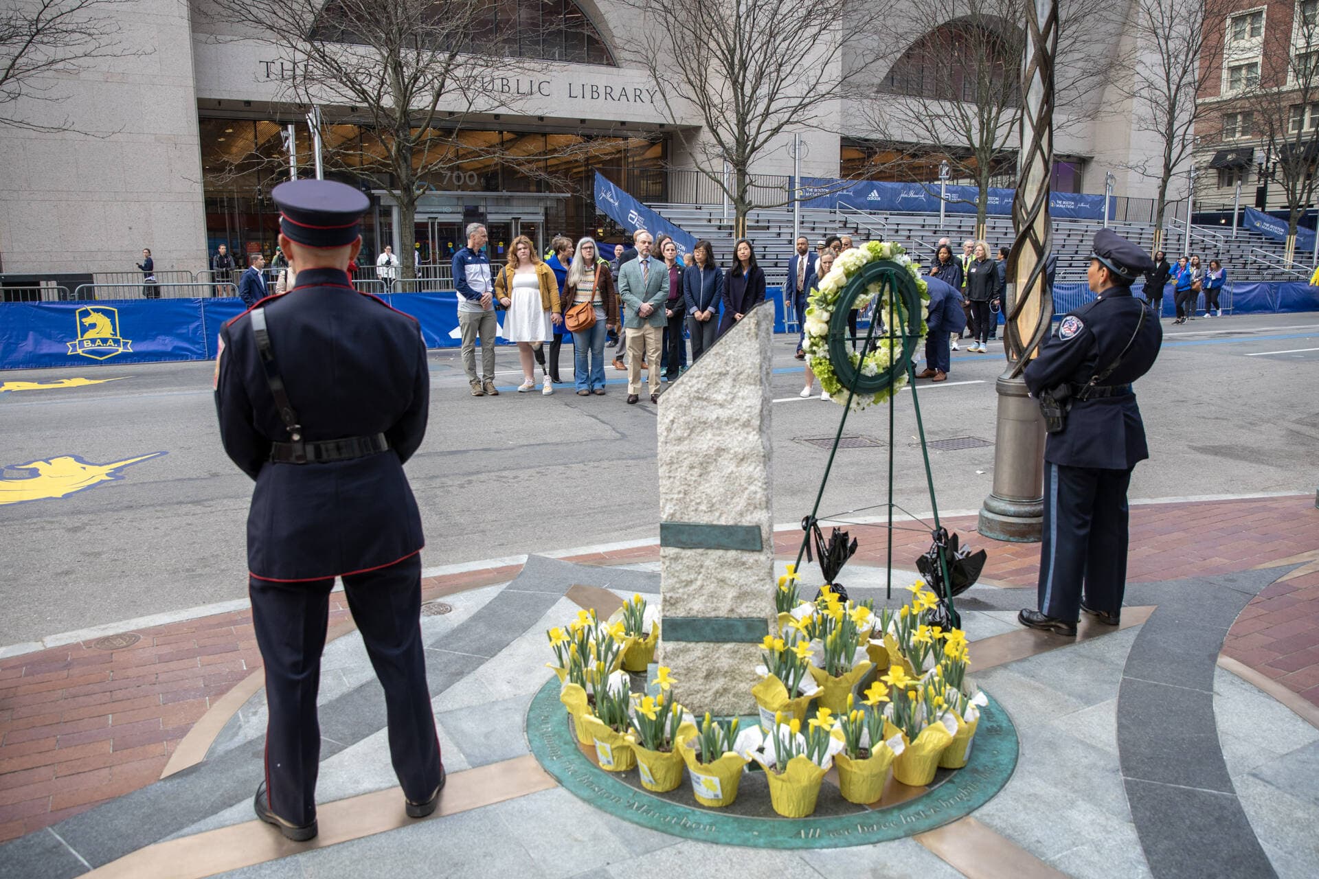 A ring of yellow daffodils and a white wreath surround a memorial for victims of the bombings. Healey and Wu stand alongside several family members who lost loved ones in the 2013 attack. (Robin Lubbock/WBUR)