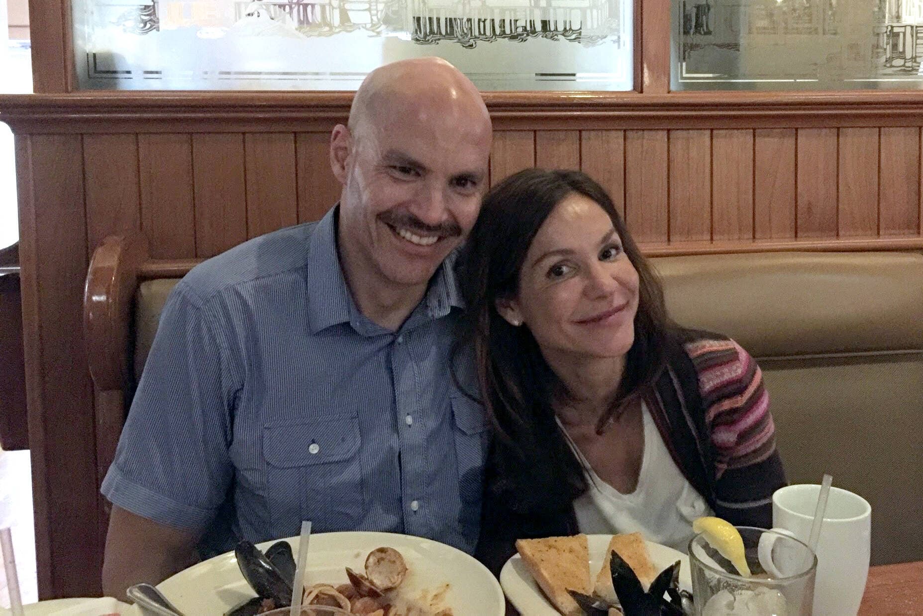 Wideman, a multiracial man, wearing a blue short-sleeved shirt, has his arm around Marta DeSoto, a Spanish woman, inside a restaurant.