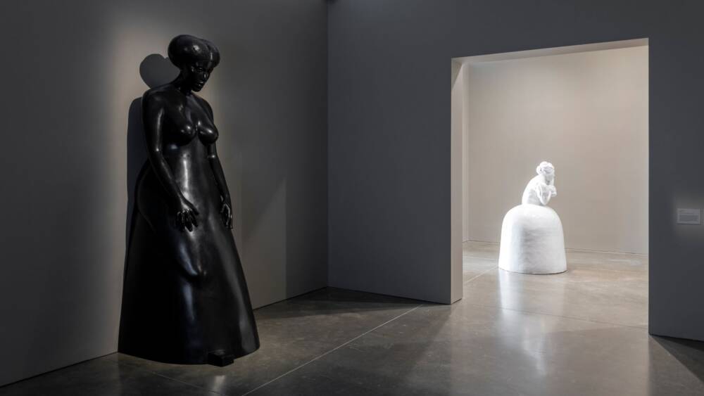A bronze sculpture of a woman stands in the forefront. A white ceramic sculpture is in the background.