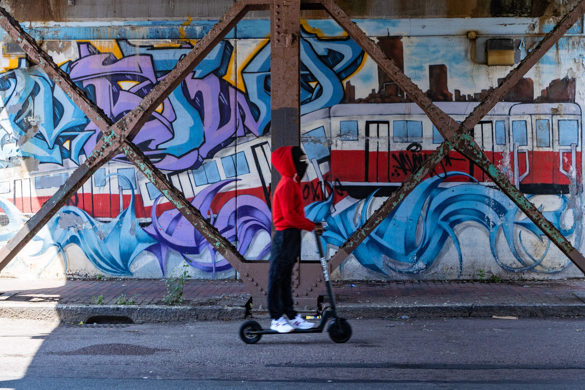 A young person rides an electric scooter beneath the Fields Corner MBTA train bridge on Dorchester Avenue. (Jesse Costa/WBUR)