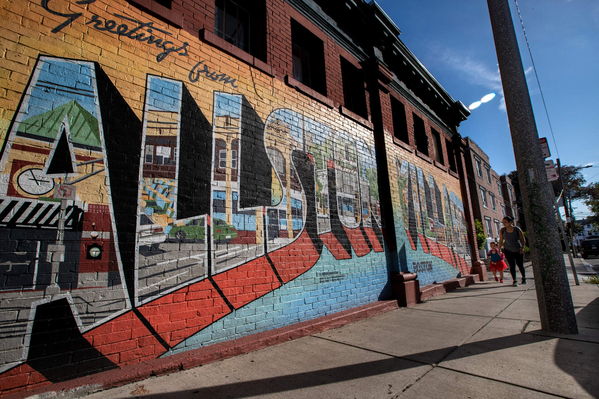 A mother and her daughter walk past the “Greetings from Allston Village” mural on Farrington Street. (Jesse Costa/WBUR)