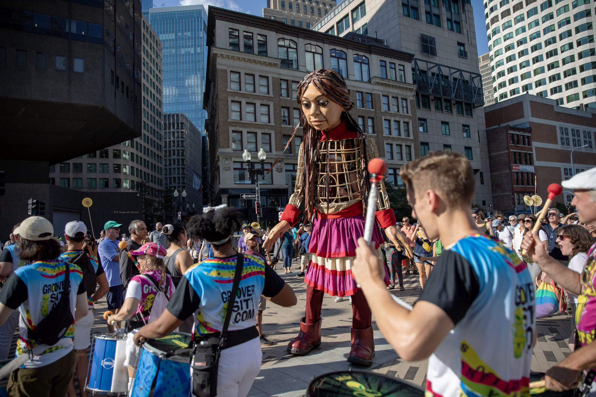 Drummers join Little Amal as she walks along Atlantic Avenue in Boston. (Robin Lubbock/WBUR)