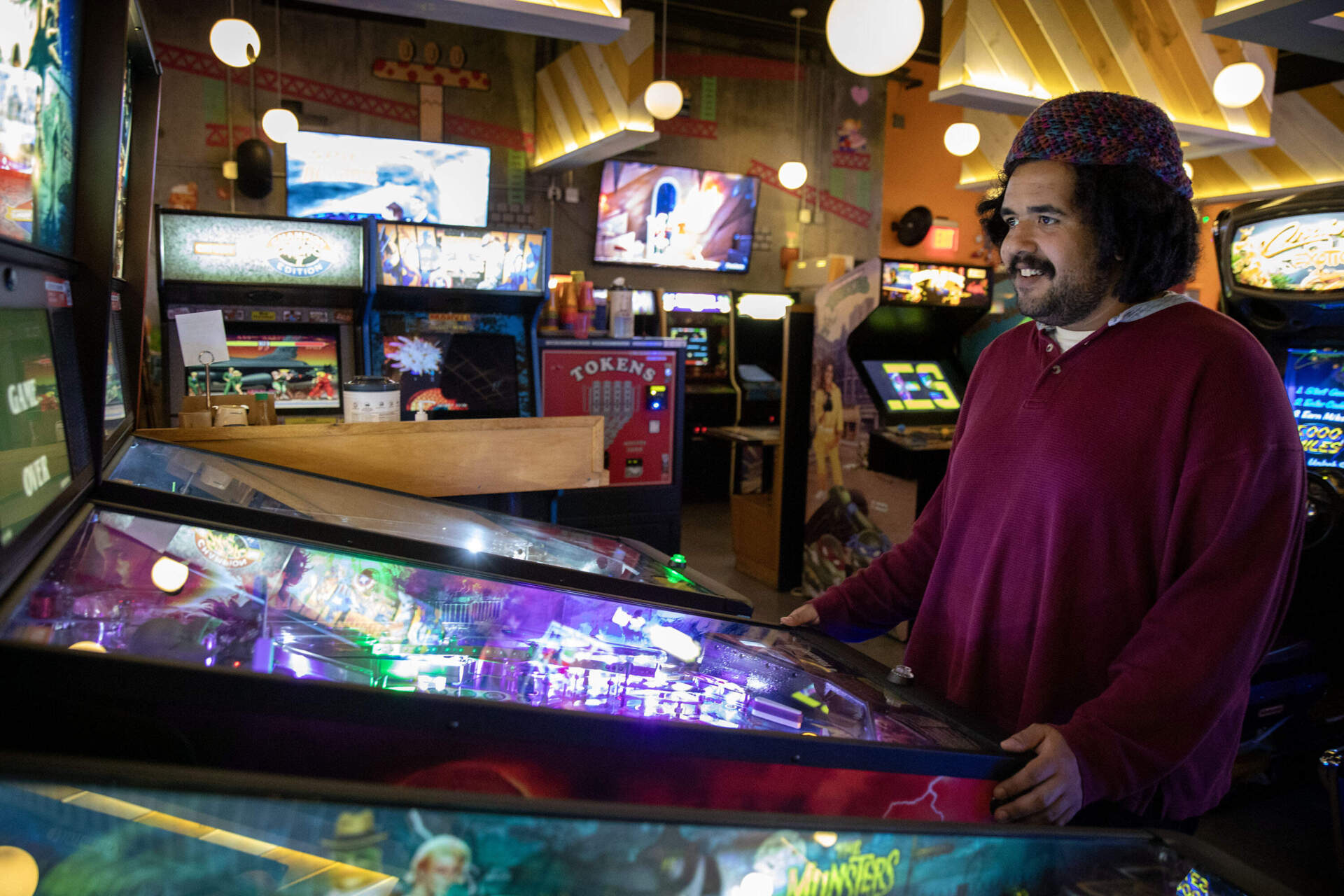 Smiling man in a burgundy-colored shirt stands at the foot of an arcade game.