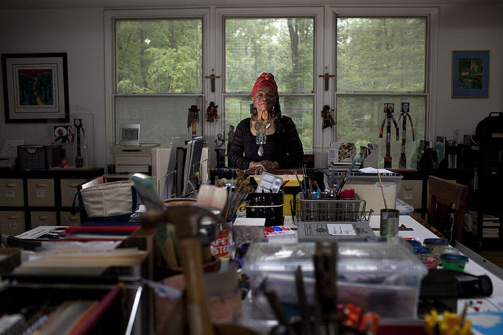 Artist Faith Ringgold stands at the head of a long table covered in art materials.