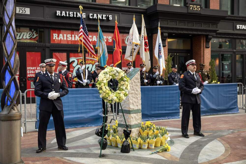 an honor guard stands in remembrance by the memorial at the site of the first bomb on Boylston Street. The memorial is surround by potted daffodils. (Robin Lubbock/WBUR)