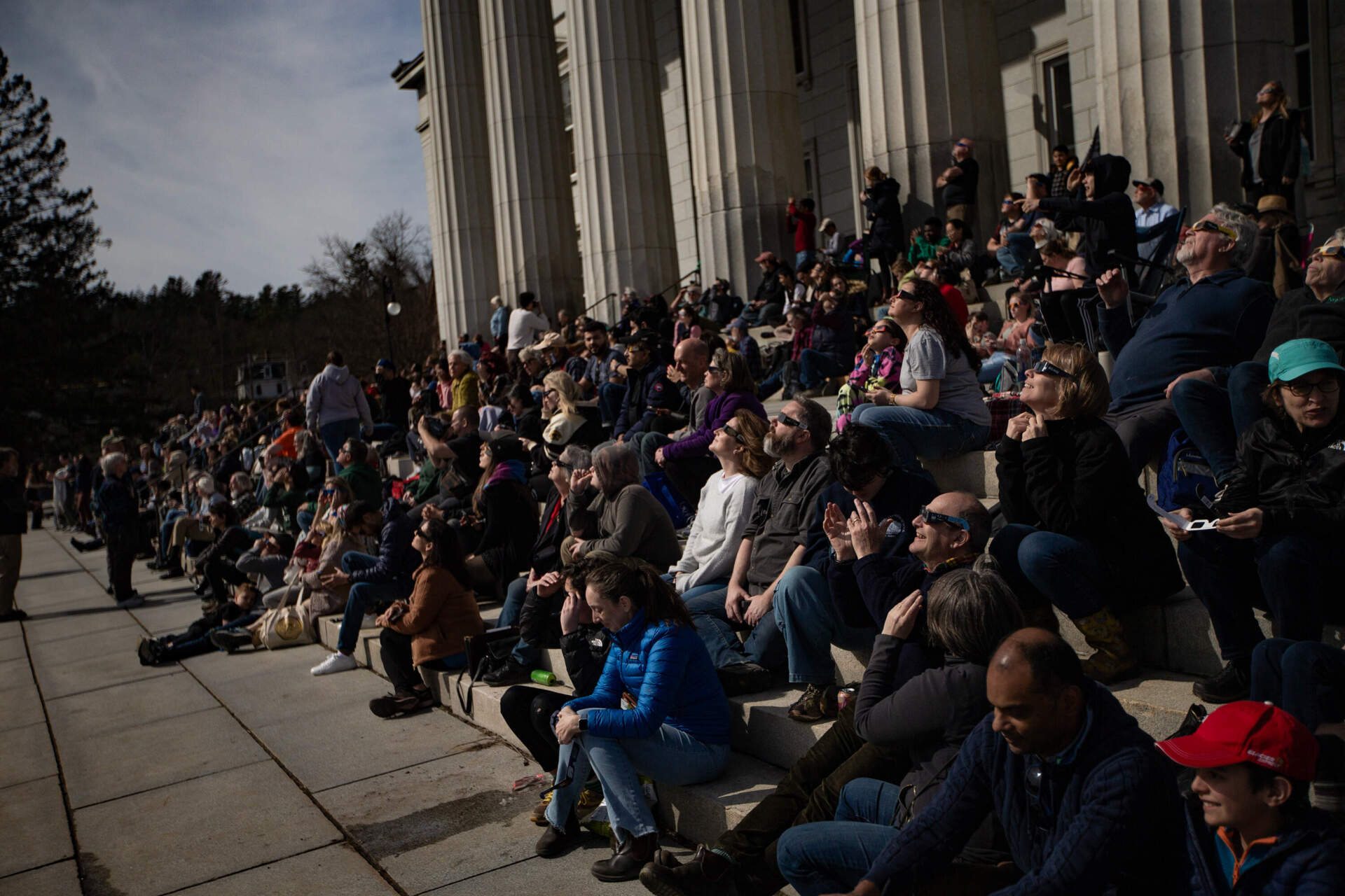 People sitting on the Vermont State House steps marvel as the moon covers the sun as the eclipse gets closer to totality as the sky darkens. (Jesse Costa/WBUR)