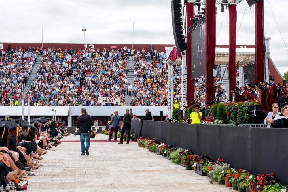 UMass Amherst Chancellor Javier Reyes stands at the podium, right, while protesters exit McGuirk Stadium during the commencement ceremony, May 18, 2024. (Ben James/NEPM)