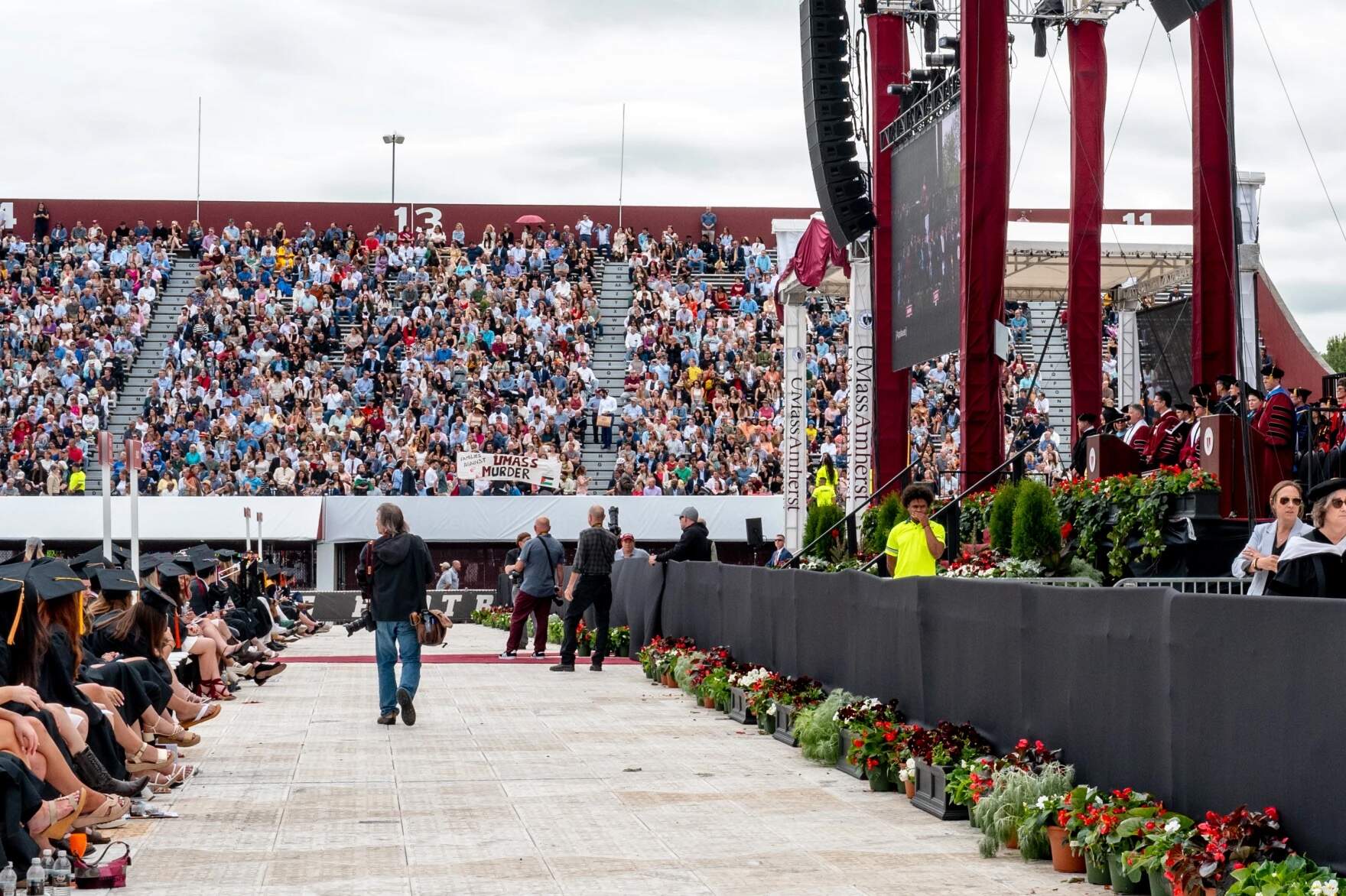 UMass Amherst Chancellor Javier Reyes stands at the podium, right, while protesters exit McGuirk Stadium during the commencement ceremony, May 18, 2024. (Ben James/NEPM)