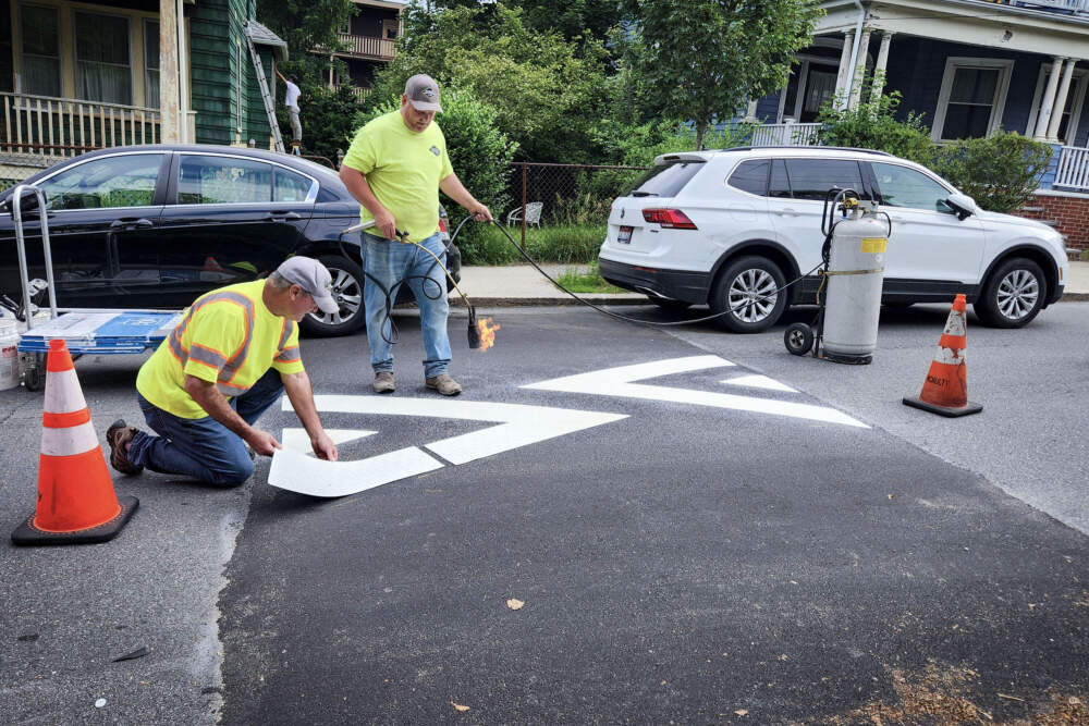 A crew works to adhere markings onto a speed hump on Peter Parley road in Jamaica Plain. One man places chevron-shaped decals on the hump (left) while another adheres the decal using a blowtorch. 