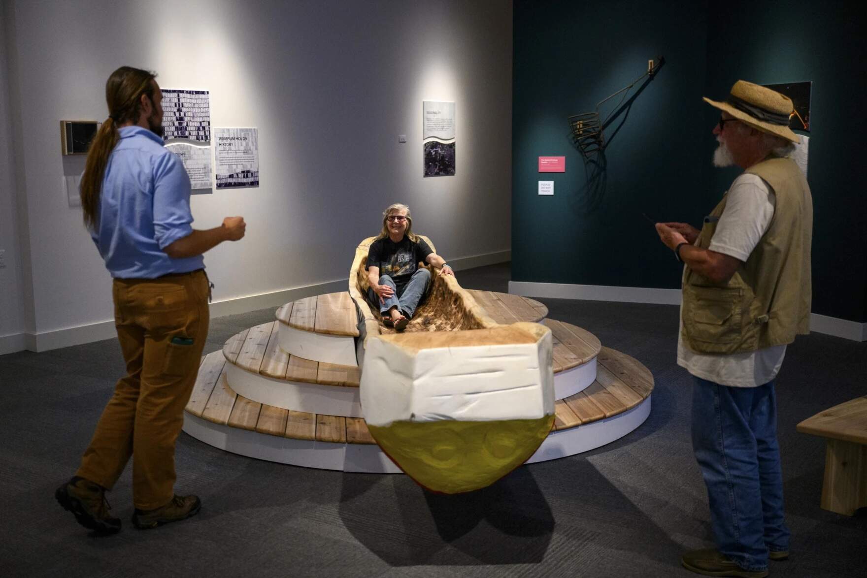 Museum visitor Margaret Sneddon sits in a 'muhshoon' as she and her husband Bill talk to museum interpreter James Weitlauf. The dugout canoe was created for the 'Entwined' exhibition using a burning method common to both West African and Native American cultures. (Ryan Caron King/Connecticut Public)