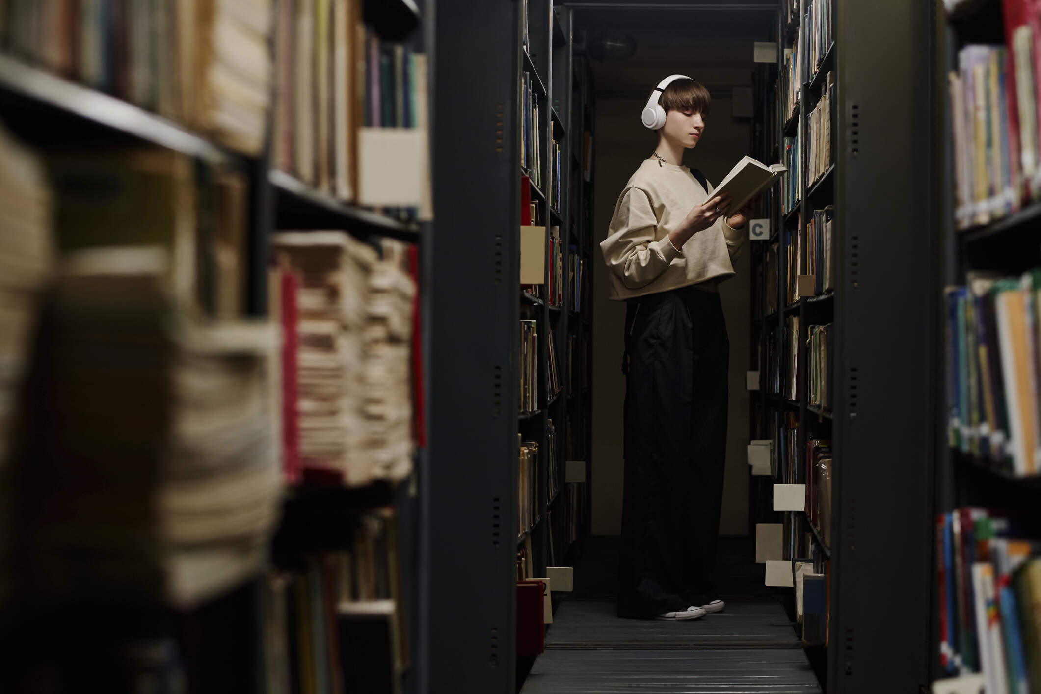 Student wearing wireless headphones and reading book near shelves in library. (Getty Images)