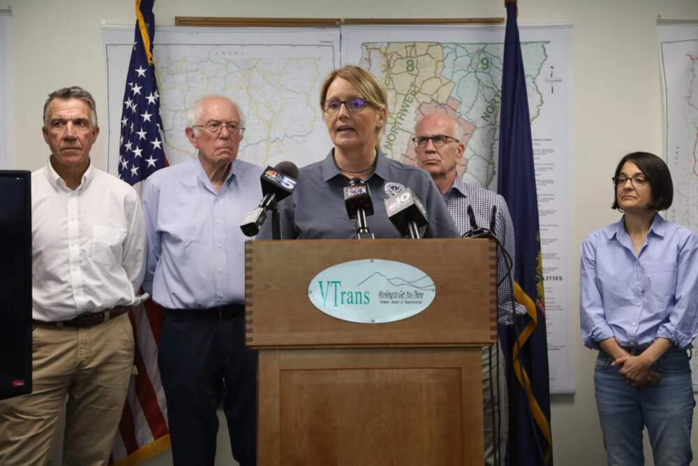Gov. Phil Scott, far left, and FEMA Administrator Deanne Criswell, at podium, discuss Vermont's flood response last July. (Mike Dougherty/Vermont Public)