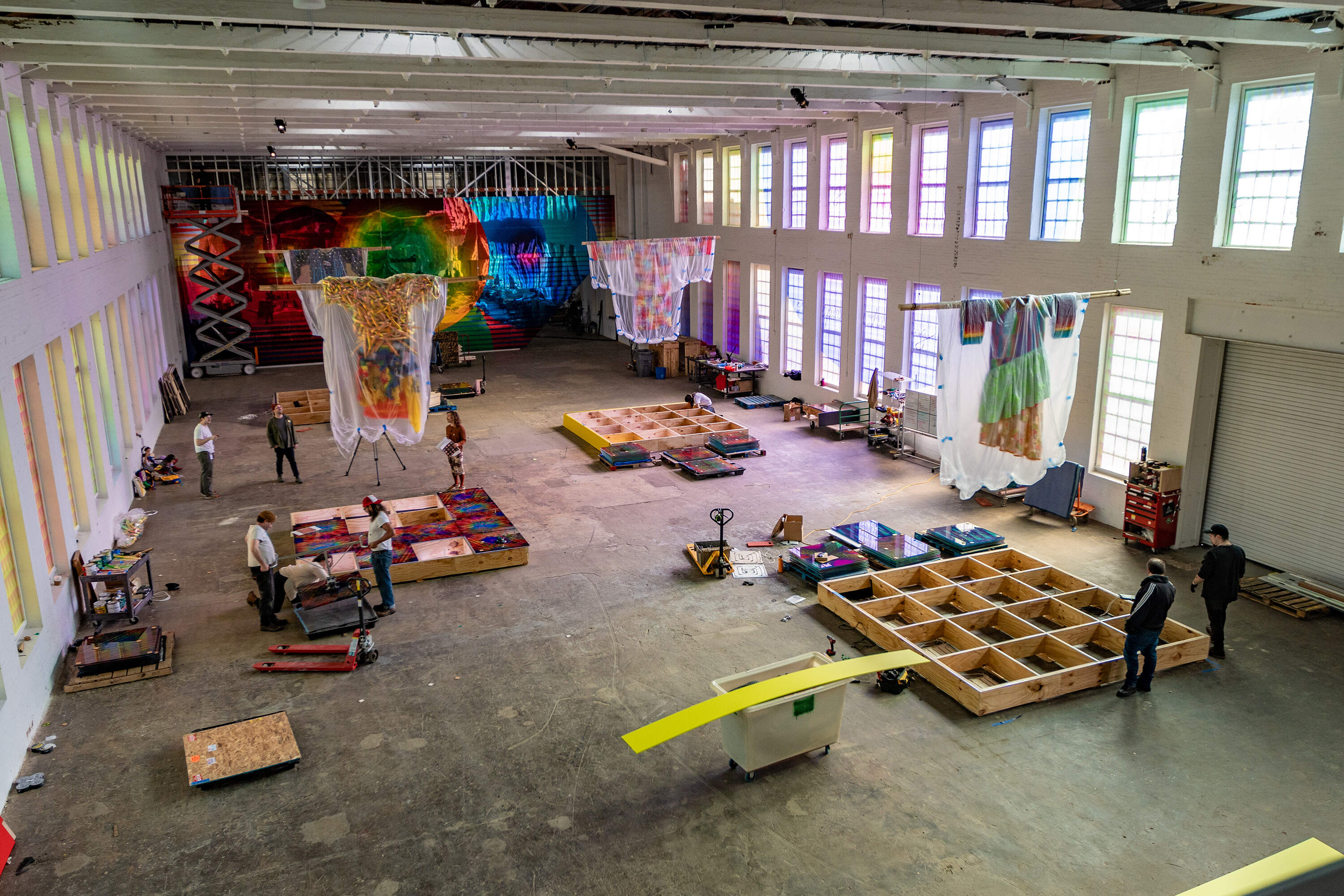 MASS MoCA staff hang Jeffrey Gibson's oversized clothing from the ceiling above 7 12×12 foot fused glass performance stages with graphic geometric patterns. (Jesse Costa/WBUR)