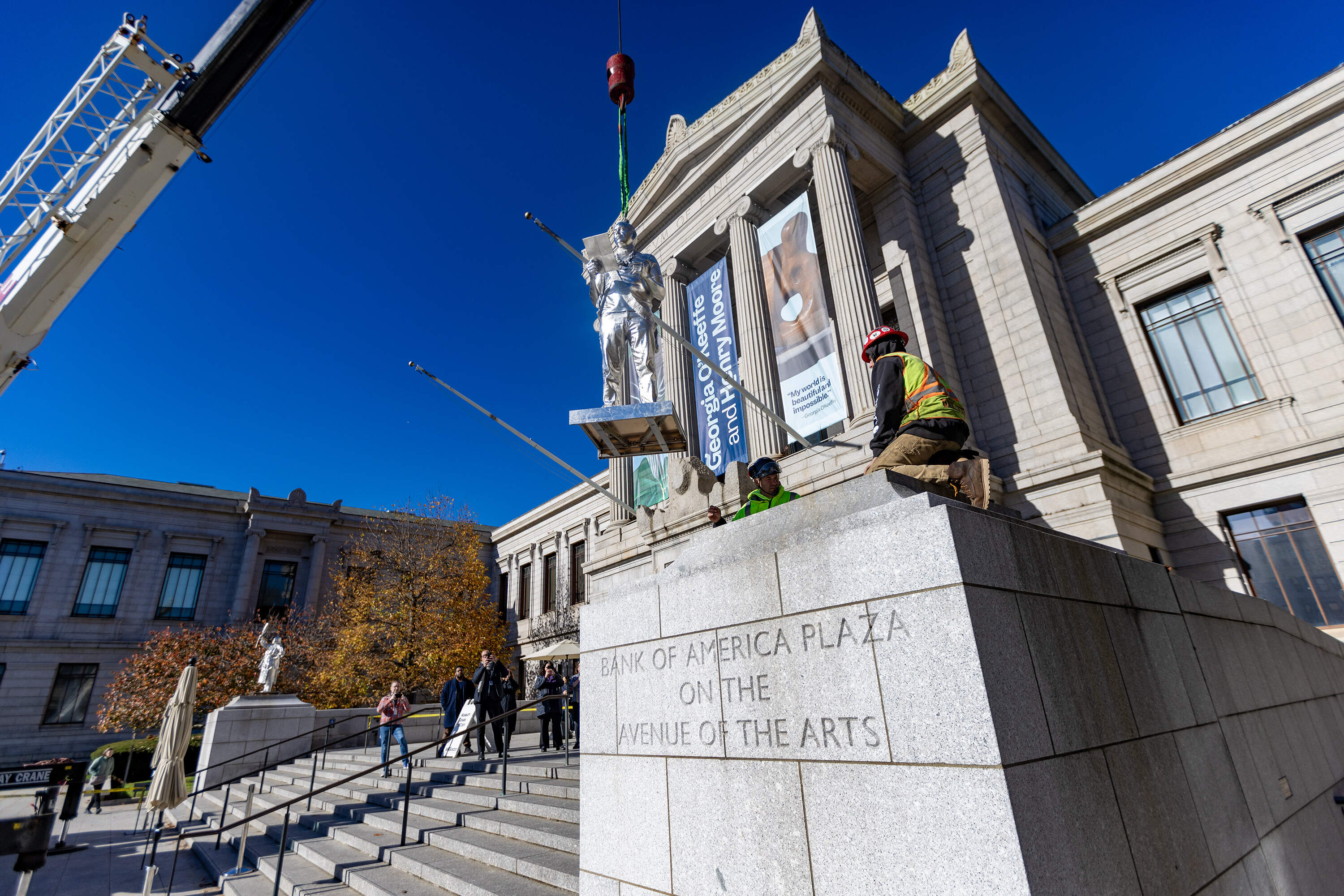 The statue of Nipmuc descendant Andre StrongBearHeart Gaines, Jr. being lowered onto one of the two large plinths after the statue of Aquinnah Wampanoag member and artist Julia Marden was installed outside the Museum of Fine Arts main entrance, part of The Knowledge Keepers by Alan Michelson. (Jesse Costa/WBUR)