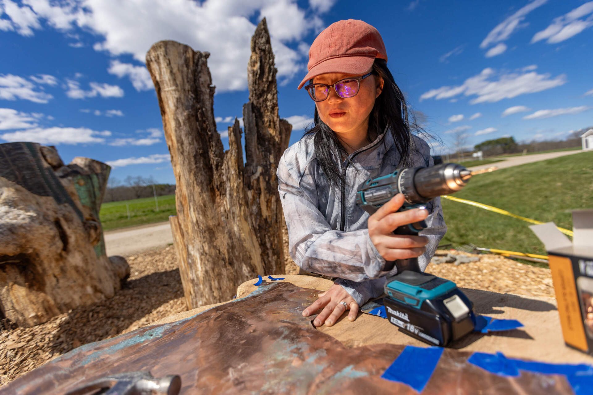 Artist Jean Shin’s face glows with the reflection of the sun off of the copper she is attaching it onto a tree stump as part of her exhibit at Appleton Farms, “Perch.”
