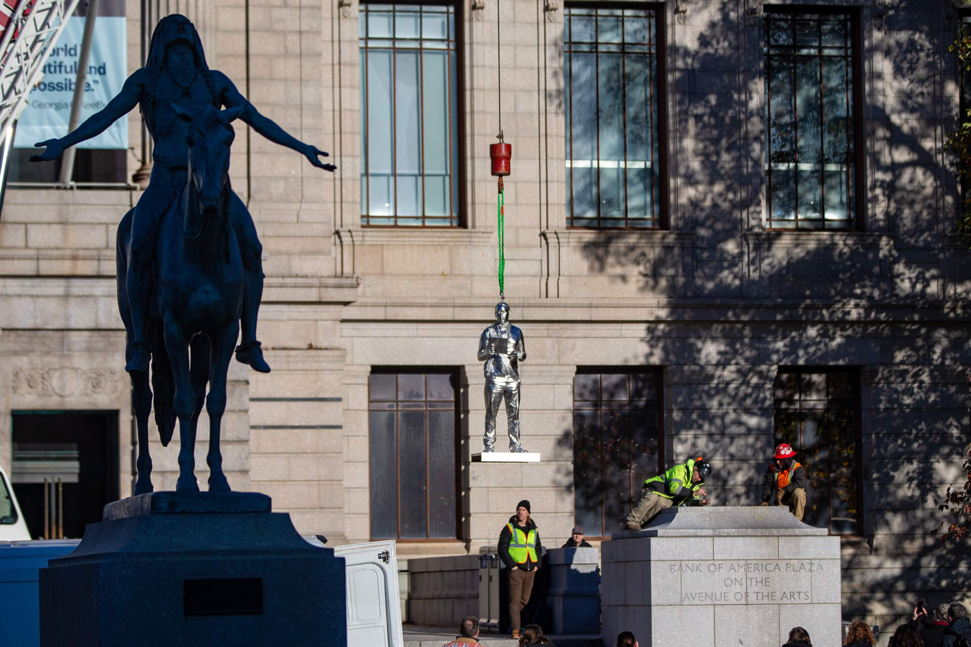 Appeal to the Great Spirit (1909), a sculpture by Cyrus Dallin, sits in the foreground while the statue of Nipmuc descendant Andre StrongBearHeart Gaines, Jr. being raised to one of the two large plinths outside the Museum of Fine Arts main entrance, part of The Knowledge Keepers by Alan Michelson.