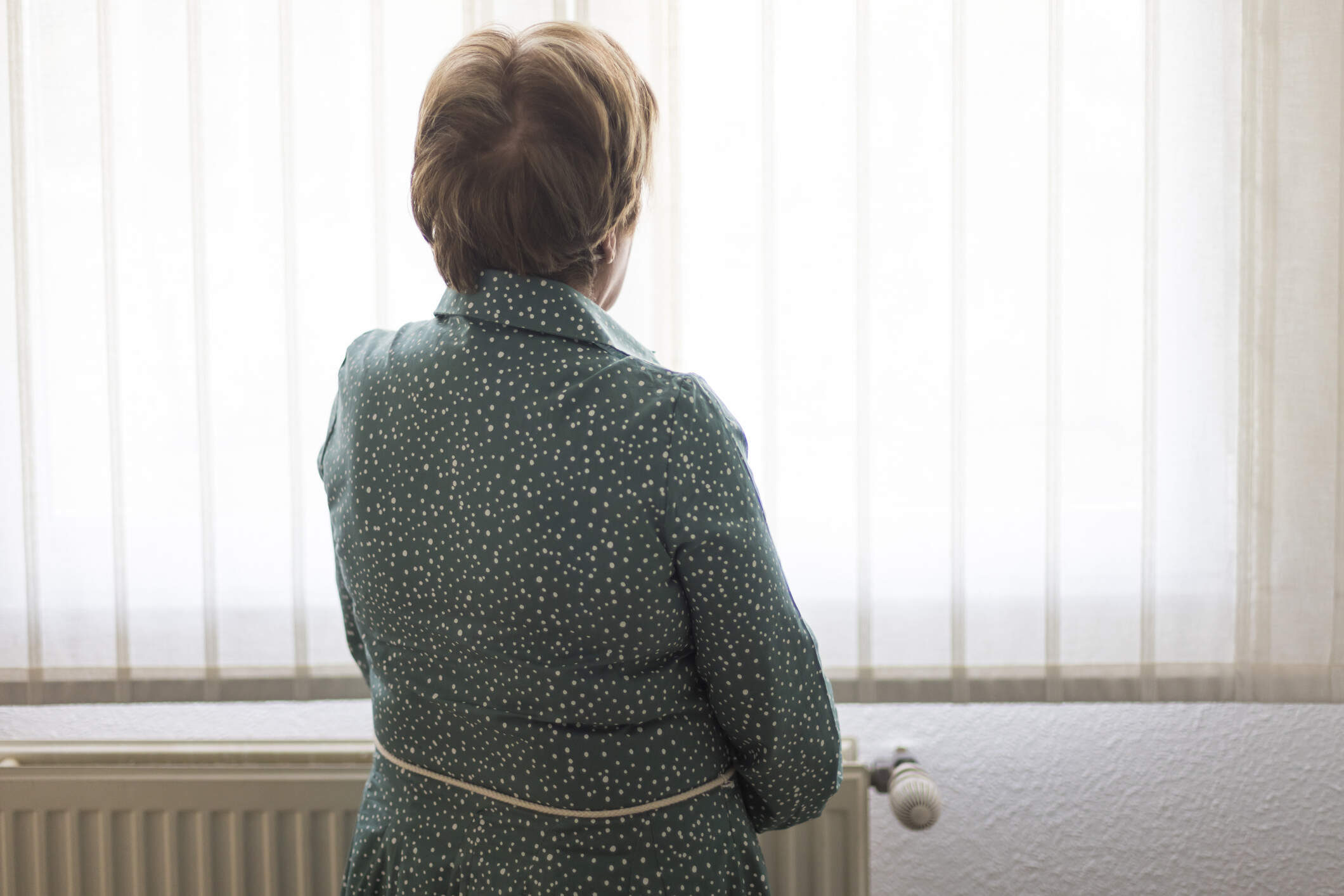 A senior woman with her back turned, looks out a window. (Getty Images)