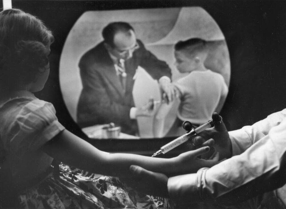 As an 8-year-old girl receives a polio vaccine, she watches a closed circuit television broadcast (showing Dr. Jonas Salk as he inoculates a boy) of a training telecast for physicians and scientists, New York, New York, April 12, 1955. (PhotoQuest/Getty Images)