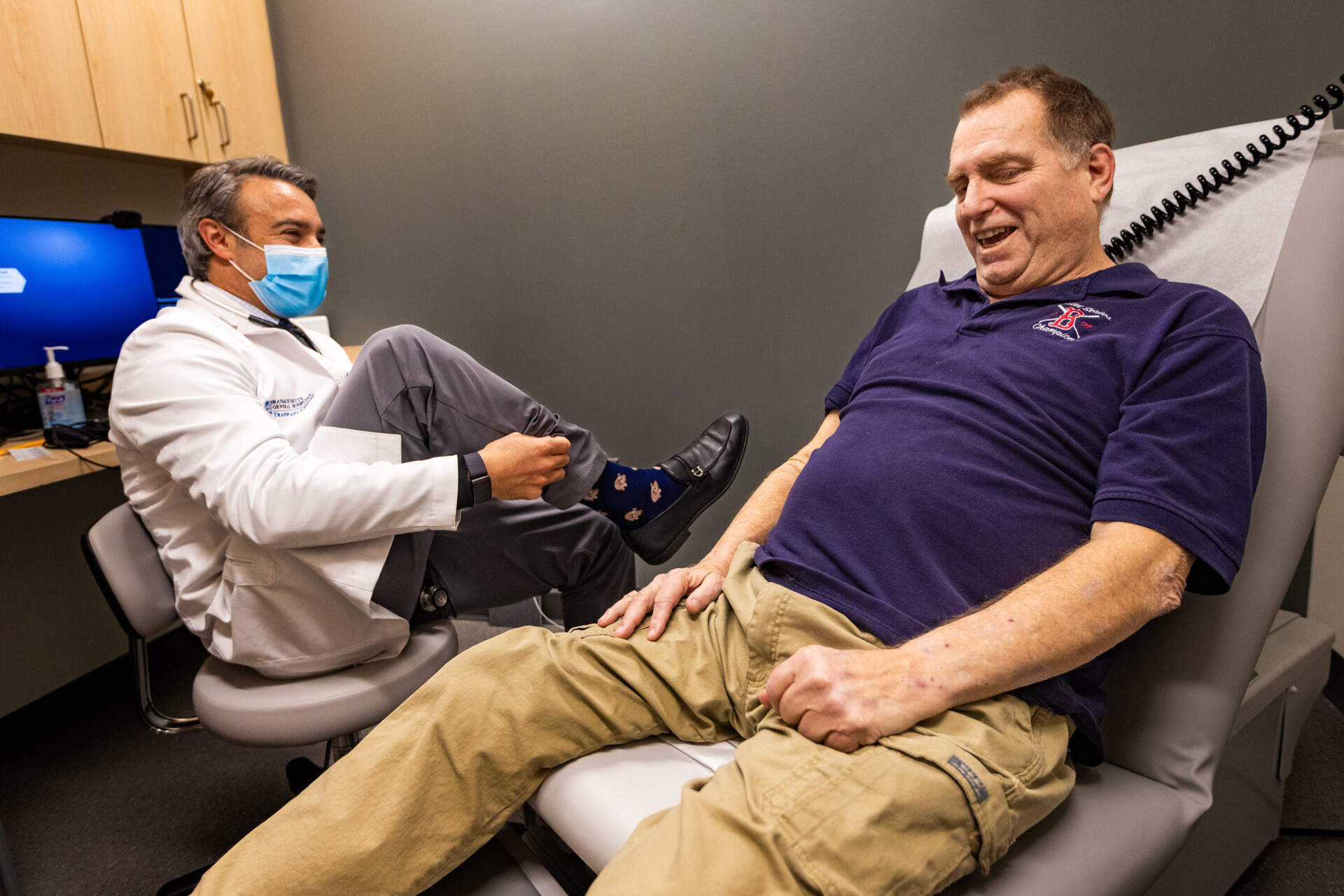 Dr. Leonardo Riella shows pig kidney recipient Tim Andrews the pig socks he is wearing at the start of Andrews' checkup at Mass General in Boston. (Jesse Costa/WBUR)