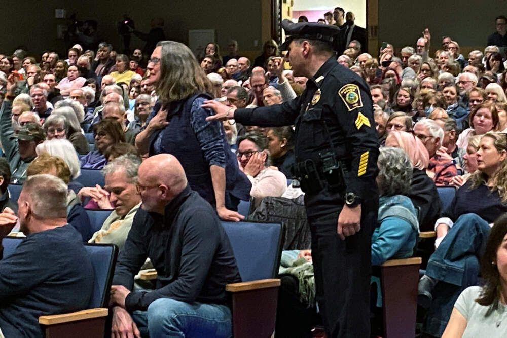 An officer escorts a woman out of the town hall after she confronted Rep. Seth Moulton about his previous remarks on transgender women in sports. (Deborah Becker/WBUR)