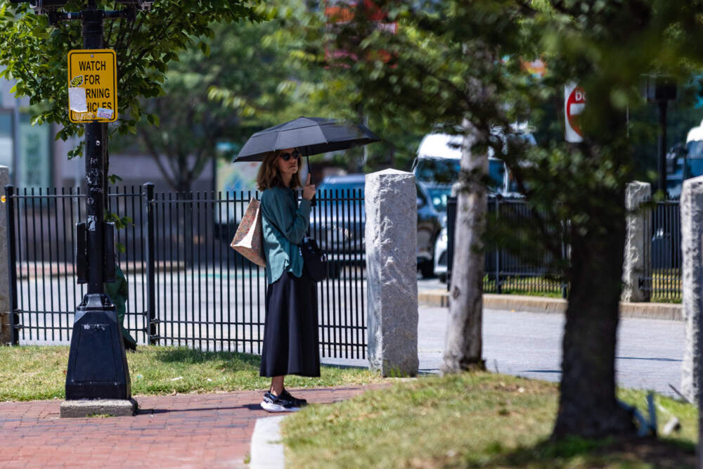 A woman with an umbrella to shield herself from the sun waits to cross Commonwealth Avenue in Kenmore Square.