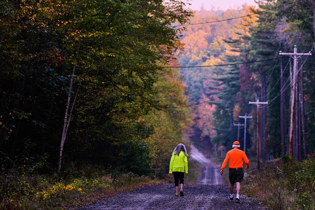 A couple, wearing protective high-visibility clothing due to hunting season, take a walk at dawn, as trees of in the distance turn to fall foliage colors, Tuesday, Oct. 7, 2025, in Chester, N.H. (Charles Krupa/AP)