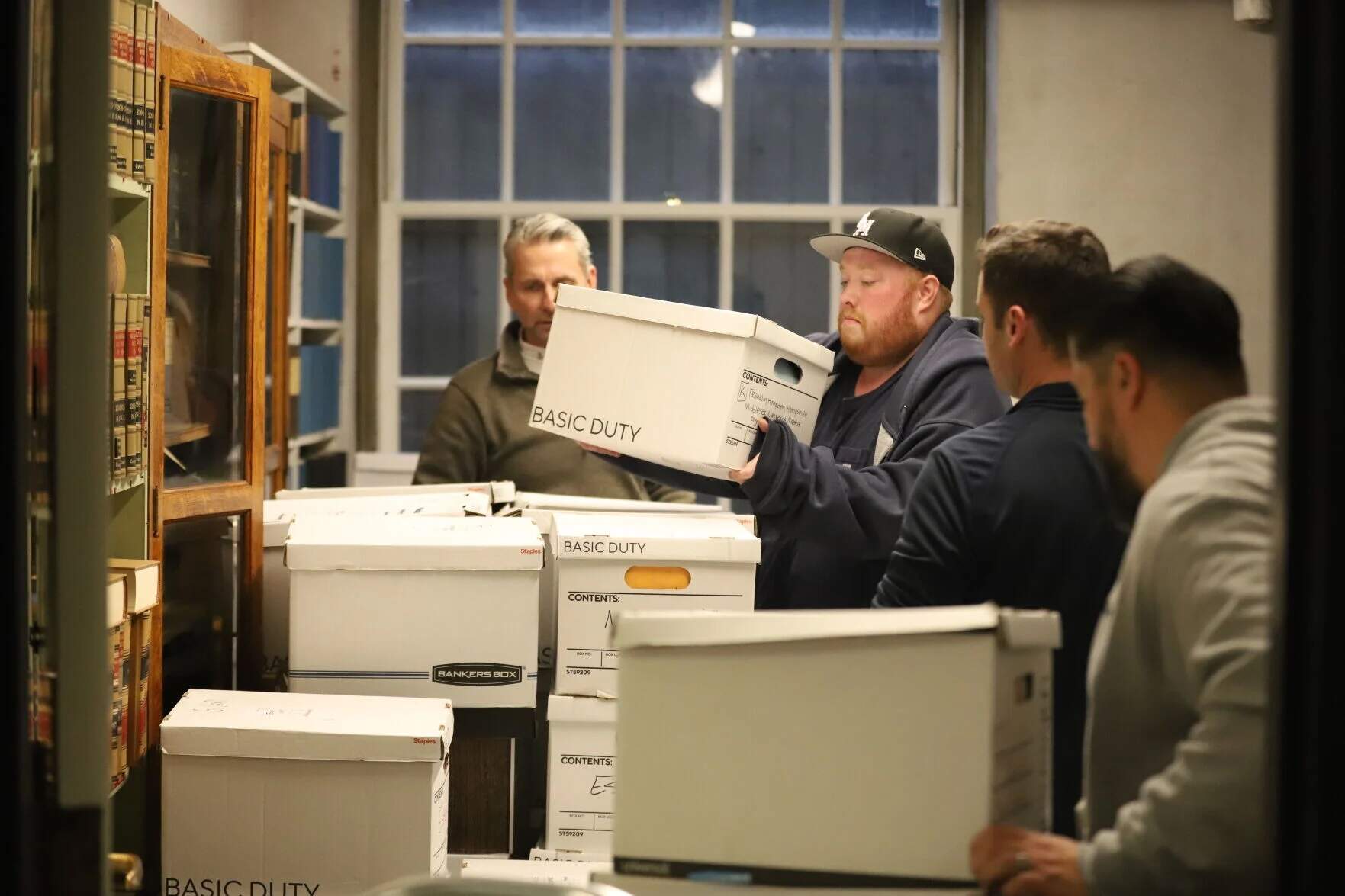 Several members of Secretary William Galvin's office and House Clerk Timothy Carroll (far left) lift boxes full of initiative petitions from dollies into Room 59 in the State House basement on Jan. 6, 2026. (Ella Adams / SHNS)