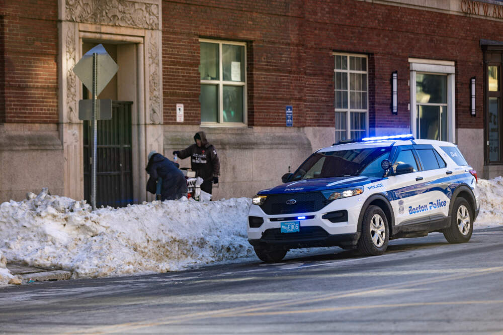 A Boston police cruiser is parked with lights flashing as two people organize their belongings in a cart in front of the entrance of AHOPE Boston on Albany Street. (Jesse Costa/WBUR)