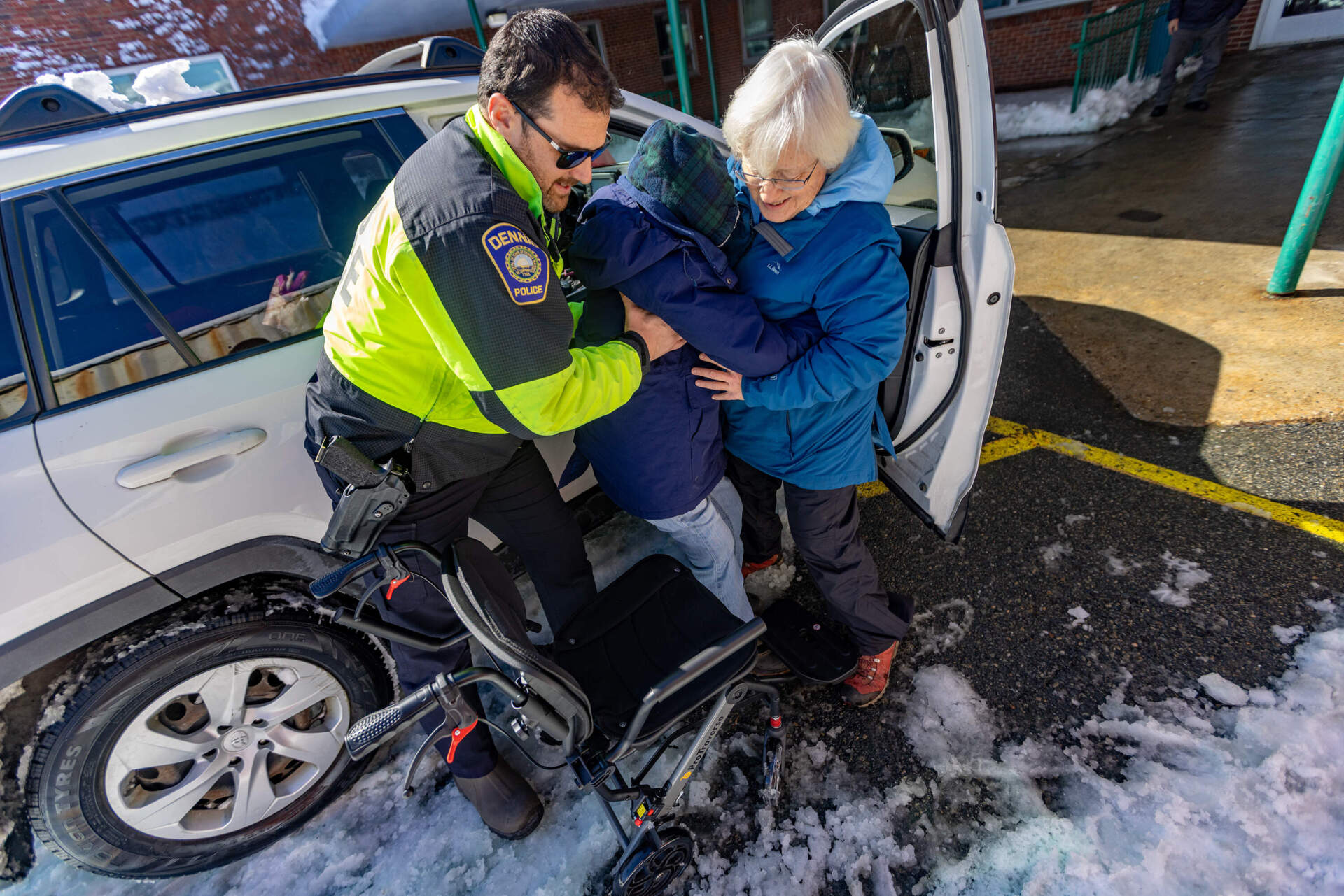 A Dennis police officer helps Barbara Cormier lift her 91-year-old mother into a wheelchair as they arrive at the emergency shelter set up at the Dennis Yarmouth Regional High School for locals without heat or power. (Jesse Costa/WBUR)