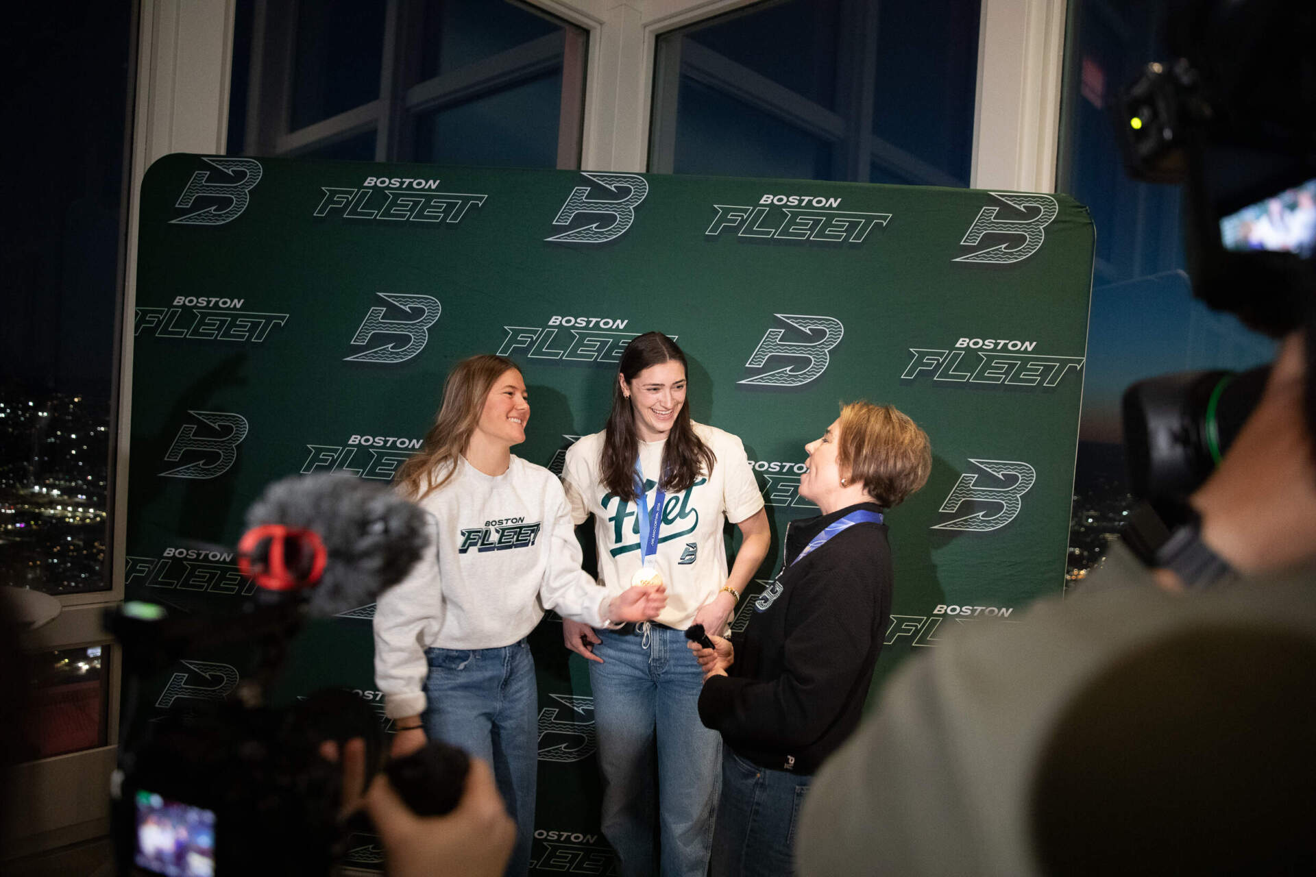 Boston Fleet defender Haley Winn (left), standing next to Boston Fleet Captain Megan Keller, lets Governor Healey try on her gold medal. (Artemisia Luk/WBUR)