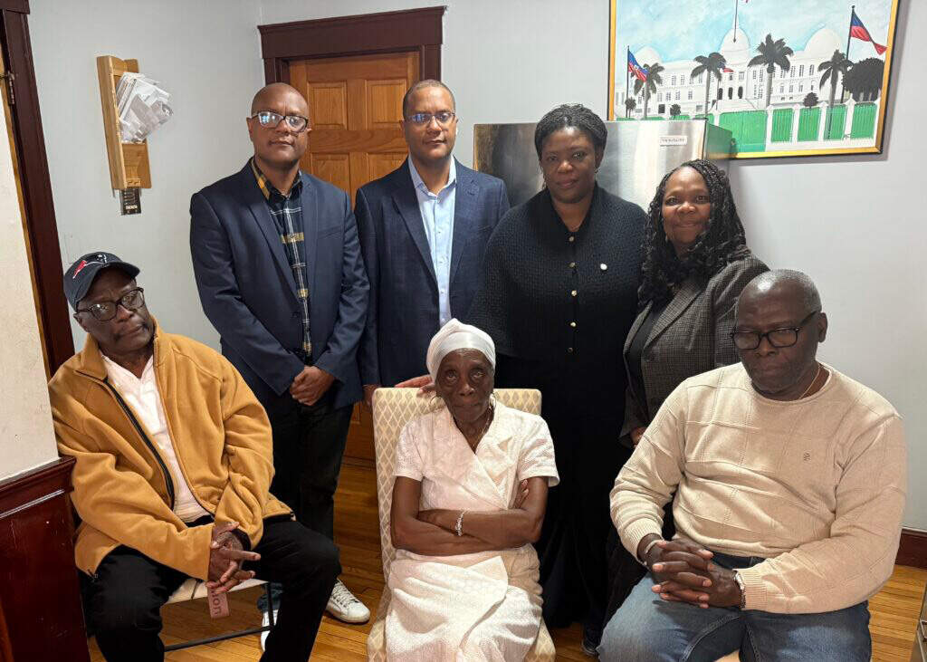 Family members of Emmanuel Damas in their home with friends and community leaders. Front, Yvon Verdier, Damas’s mother Edele-Cia Nelson, and Joseph Verdier. Back, Presner Nelson, Presly Nelson, City Councillor Ruthzee Louijeune, and IFSI Director Dr. Geralde Gabeau. (Courtesy photo via Dorchester Reporter)
