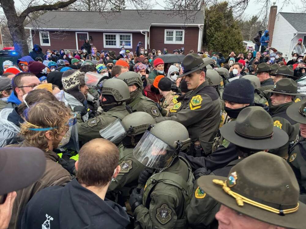 Vermont State Police in tactical gear face off against a crowd of protesters blocking the departure of Immigration and Customs Enforcement vehicles outside a South Burlington house. (Zoe McDonald/Vermont Public)